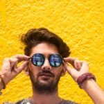 Young man with sunglasses standing against a vibrant yellow wall in daylight.
